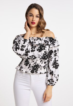 Vrouw met golvend haar, die rode lippenstift draagt, een witte off-shoulder blouse met zwart bloemmotief en witte broek, poseert met haar hand bij haar gezicht.