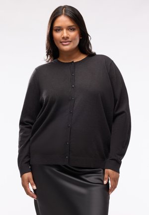 Donna con capelli castani di media lunghezza che indossa un cardigan nero con bottoni e una gonna nera lucida, in piedi davanti a uno sfondo chiaro e semplice.