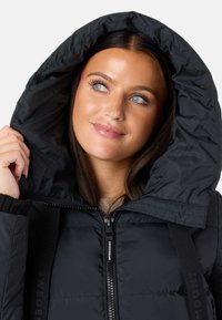 Giacca a vento nera imbottita con un grande cappuccio regolabile, zip frontale e polsini a coste. Presenta un branding sottile sulle cinghie e una texture liscia.