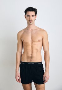 Črne bokserice Calvin Klein z elastičnim pasom, ki ima blagovno znamko. Model ima golo torzo in nevtralno ozadje.
