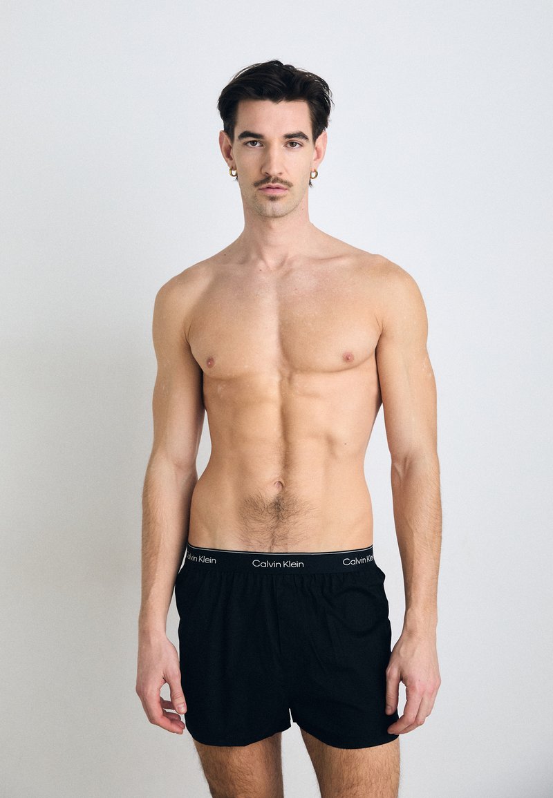 Črne bokserice Calvin Klein z elastičnim pasom, ki ima blagovno znamko. Model ima golo torzo in nevtralno ozadje.
