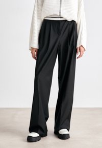 Schwarze, weit geschnittene Hosen aus glattem Stoff mit hohem Bund; kombiniert mit klobigen, schwarz-weißen Schuhen. Oben ein strukturiertes, weißes Sweatshirt.