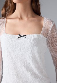 Kvinne i hvit bluse med blonde og lange ermer, med firkantet halslinje og en liten svart sløyfe midt foran.