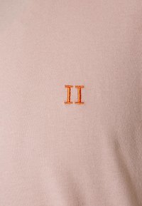 Light pink fabric with a smooth texture, featuring an orange embroidered "II" design on the left side. Simple and minimalistic aesthetic.