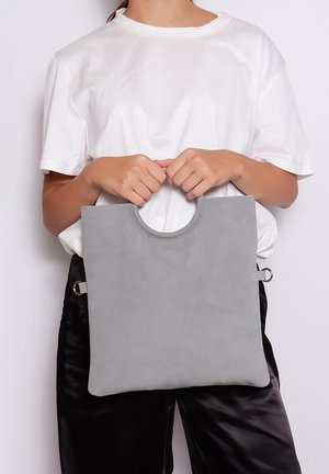 Borsa tote in suede grigia con manico circolare e anelli laterali, caratterizzata da un design minimalista. Indossata con una camicia bianca e pantaloni neri.