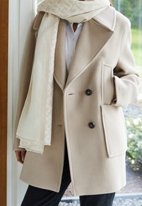 Beige jas van wolmix met reverskraag, dubbele knopen en zijzakken, gecombineerd met een lichtbeige sjaal met een patroonrand.