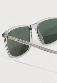 Διαφανείς γυαλιά ηλίου με πράσινους φακούς, με μεταλλικά χρυσά στοιχεία στους βραχίονες και κομψό, γωνιακό σχέδιο.