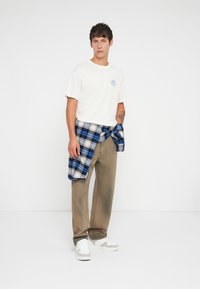 Off-white t-shirt with blue graphic, beige loose-fit pants, and a blue plaid shirt tied around the waist. White sneakers complete the look.