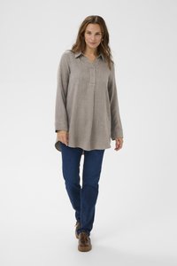 Kvinna som går framåt iklädd en lång beige skjorta, mörkblå jeans och bruna snörskor mot en enfärgad vit bakgrund.