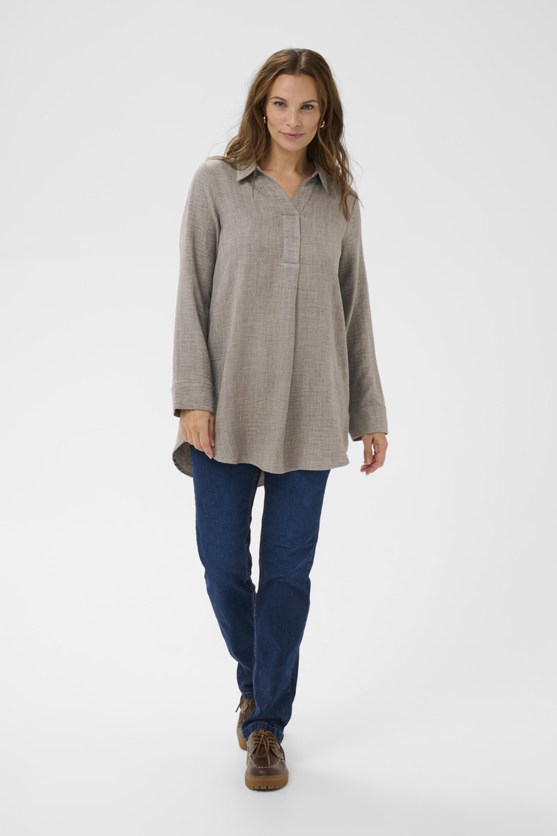 Kvinna som går framåt iklädd en lång beige skjorta, mörkblå jeans och bruna snörskor mot en enfärgad vit bakgrund.