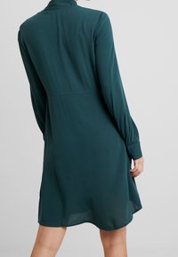 Vestido largo de manga larga en verde azulado con cuello alto, cuerpo semi-ajustado y falda acampanada. Tejido suave con un ligero brillo y aberturas laterales.