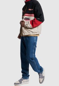 Bunda v barevném bloku v černé, červené a krémové barvě s akcenty loga, doplněná volnými modrými džínami a šedými teniskami. Texturovaná látka.