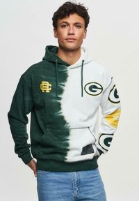 Huppari, jossa on jakautunut muotoilu; vasen puoli tummanvihreä, oikea puoli valkoinen. Siinä on vihreät ja kultaiset logot, säädettävä naru ja etutasku.