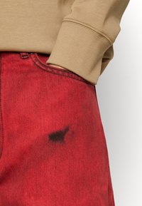 Jeans in denim rosso con una macchia nera, abbinati a una camicia a maniche lunghe beige con polsini a coste. Cuciture visibili lungo i jeans.