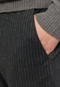 Zwarte broek met een fijne streep en een zachte textuur, voorzien van een zijzak en een grijze ribbeltrui die gedeeltelijk zichtbaar is aan de bovenrand.