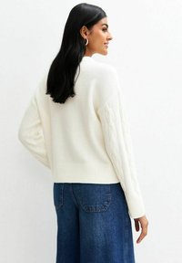 Frau mit langen schwarzen Haaren, die einen weißen, grob gestrickten Pullover und blaue Jeans trägt, seitlich blickend mit leichtem Lächeln.