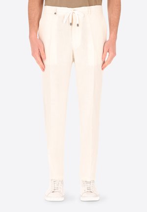 Uomo che indossa pantaloni écru con coulisse e sneakers bianche, in piedi con le mani rilassate lungo i fianchi.