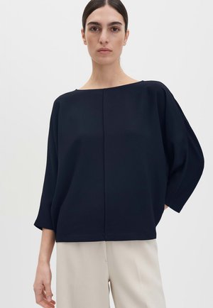 Frau mit dunklen Haaren, die ein locker sitzendes marineblaues Langarmshirt und hellbeige Hosen trägt, vor einem schlichten weißen Hintergrund stehend.