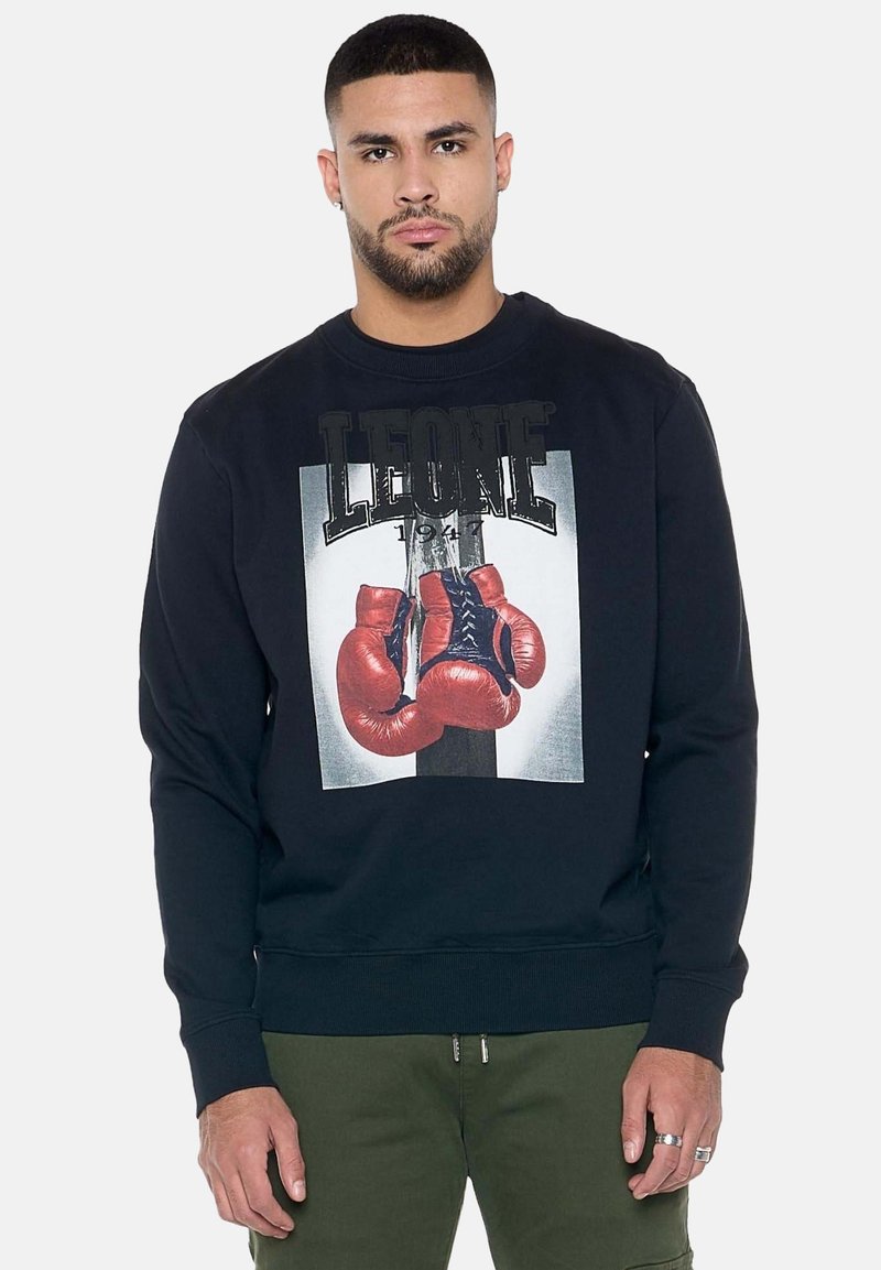 Homme aux cheveux courts et à la barbe portant un sweat-shirt noir avec des gants de boxe rouges et le texte "LEONE 1947" sur le devant.