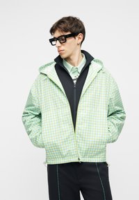 Junger Mann mit schwarzer Brille und einer hellgrünen, karierten Kapuzenjacke über einer schwarzen Reißverschlussjacke, der seitlich schaut und die Hände in den Taschen hat.