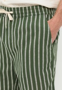 Πράσινο και λευκό κάθετο ριγέ παντελόνι με ελαστική μέση και λευκό κορδόνι, χέρι τοποθετημένο μέσα στην δεξιά τσέπη.