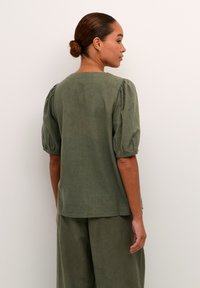 Blouse en lin vert olive avec manches bouffantes, col rond et coupe décontractée. Texture lisse, détails de couture minimaux. Design minimaliste.