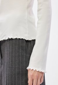 Bluză albă cu mâneci lungi și detalii cu margini desfăcute, asortată cu pantaloni gri închis cu dungi subțiri. Materialul pare moale și ușor elastic.