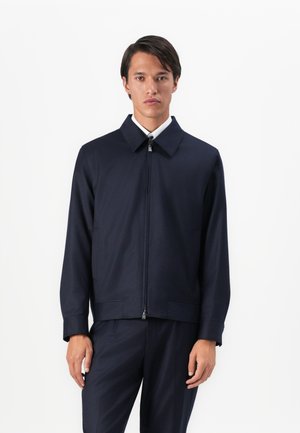 Man wearing a navy blue zip-up jacket with a collar and matching navy trousers, standing against a plain white background.