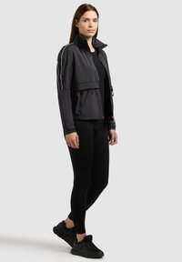 Femme debout portant une veste zippée noire avec des rayures blanches sur les manches, des leggings noirs et des baskets noires sur un fond uni.