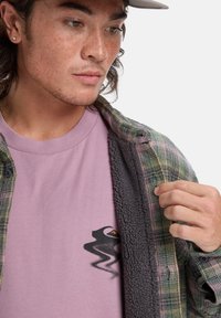 Karierte Flanellhemd in Grün und Pink über einem lavendelfarbenen T-Shirt mit schwarzem Motiv. Weiche Textur mit sichtbarem Fleecefutter.