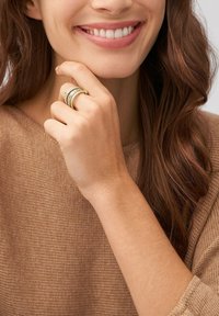 Bague dorée avec plusieurs bandes aux finitions texturées et lisses, portée sur une main à la peau claire devant un fond de pull marron.