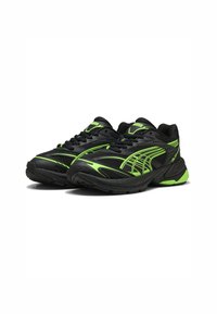 Zapatos deportivos negros con acentos verdes, con parte superior de malla, suela texturizada y líneas de diseño distintivas. Cierre con cordones.
