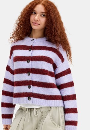 Jonge vrouw met krullend rood haar die een paarse en donkerrode gestreepte trui met knopen draagt en lichtbeige broek tegen een effen achtergrond.