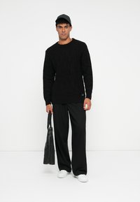 Schwarzer Pullover mit Kabelstrickmuster und langen Ärmeln, kombiniert mit schwarzen weiten Hosen. Weiße Sneakers und eine dunkle Mütze, während eine schwarze Tasche gehalten wird.
