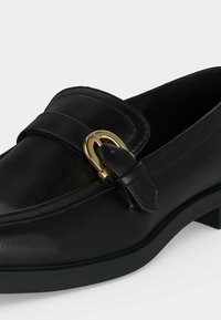 Mocassin en cuir noir avec un détail de boucle dorée sur la bride et un petit talon empilé sur fond blanc.