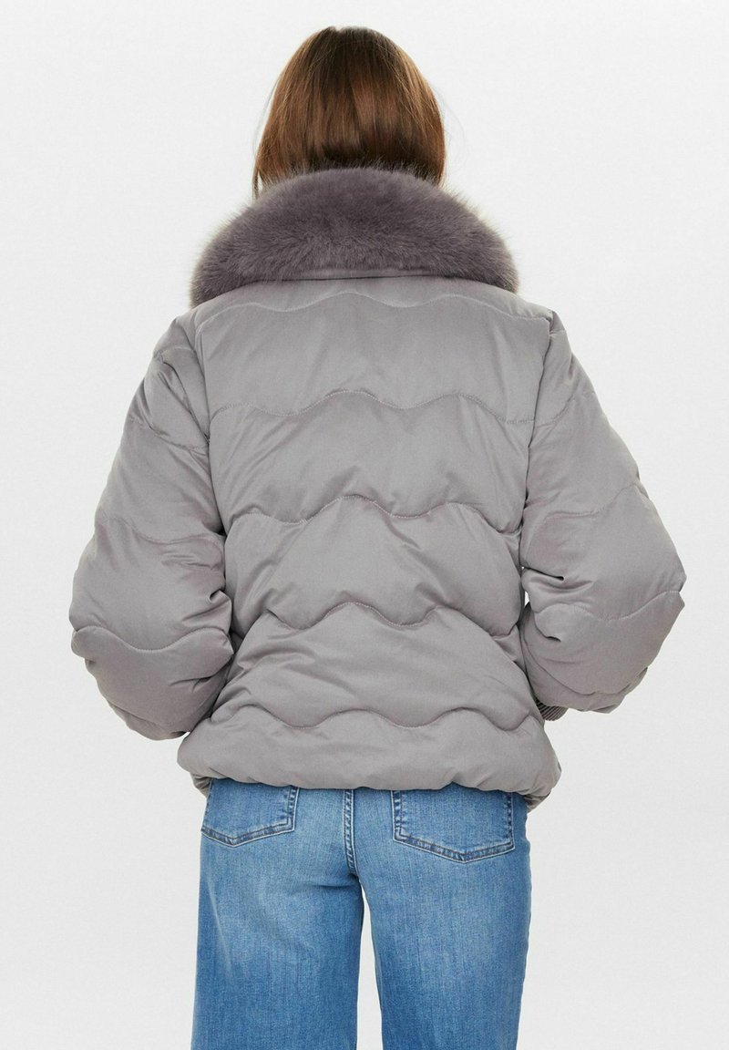 Graue Daunenjacke mit einem breiten, plüschigen Pelzkragen, gepolstertem Design mit wellenförmiger Naht und elastischen Bündchen. Kombiniert mit blauen Jeans.