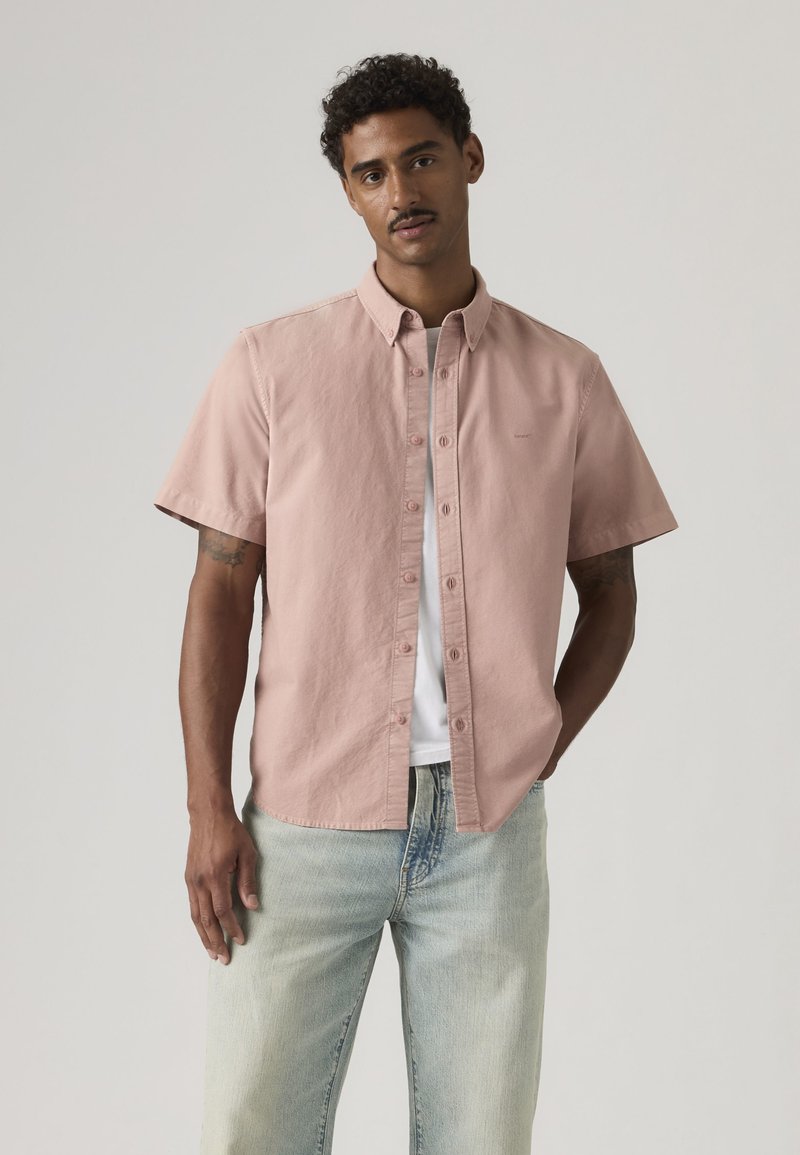 Mann trägt ein hellrosa kurzärmeliges Hemd über einem weißen T-Shirt und hellblauen Jeans, steht mit einer Hand in der Hosentasche.