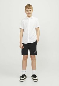 Biała koszulka z krótkim rękawem i czarne szorty z logo; czarne sneakersy z białymi akcentami, w zestawieniu z białymi skarpetkami. Prosty, casualowy strój.
