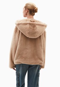 B�éžová umělá kožešinová bunda s velkou kapucí, má volný střih a měkkou texturu, kombinovaná s modrými džínovými cargo kalhotami.