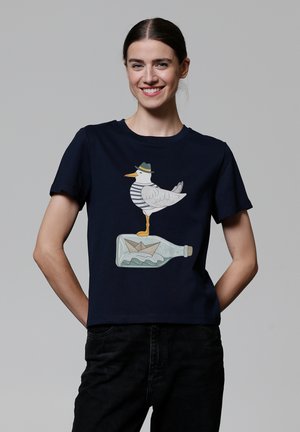 T-shirt bleu marine avec un graphique représentant une mouette portant une chemise rayée et un chapeau, se tenant sur une bouteille avec un bateau en papier à l'intérieur.