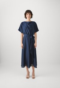 Robe midi bleu marine à manches courtes, avec une taille ceinturée et un ourlet volé. Tissu lisse avec un drapé subtil. Portée avec des sandales à talons nudes.