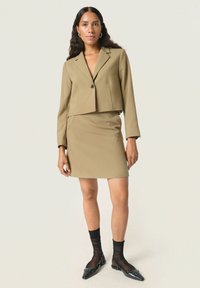 Beige kort blazer med en knapp, tillsammans med en figurnära beige kjol. Modellen bär svarta genomskinliga strumpor och svarta pumps med spetsig tå.