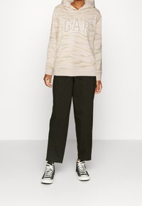 Sweat à capuche beige avec un motif zèbre subtil et le logo "GAP" blanc ; assorti à un pantalon noir ample et des baskets montantes noires.