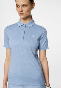 Vaaleansininen polo-paita, jossa on teksturoitu aaltoilu kuvio, viisi nappia edessä ja lyhyet hihat. Paidassa on pieni valkoinen logo rinnassa.