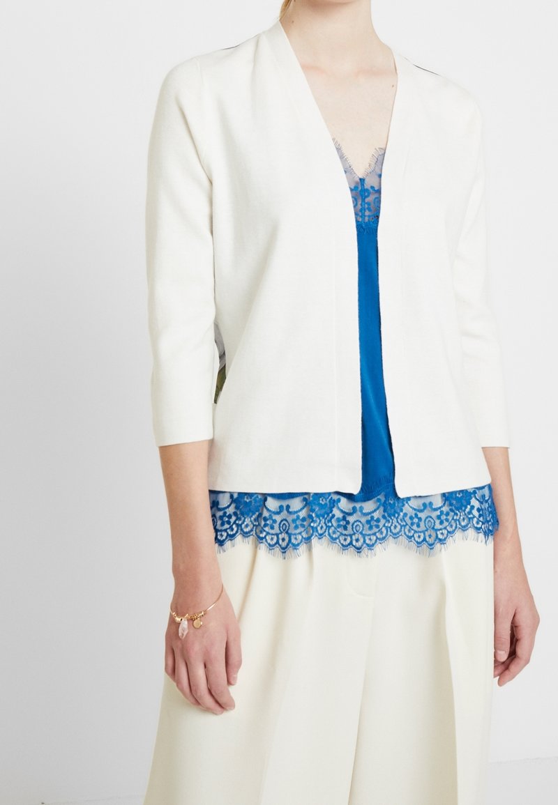 Žena v bílém kardiganu s otevřeným předním dílem přes modrou tílko s krajkovými detaily a širokými bílými kalhotami, s zlatým náramkem s přívěsky.