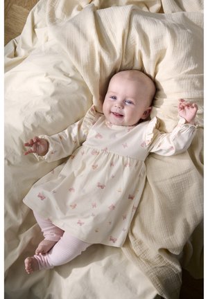 Uśmiechające się niemowlę leżące na kremowych kocach, ubrane w bladą sukienkę z nadrukiem motyli oraz różowe prążkowane legginsy.