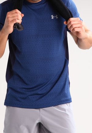 Man draagt een blauw gestructureerd Under Armour-shirt en lichtgrijze korte broek, houdt een zwarte handdoek om zijn nek vast.