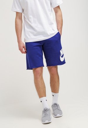 Mężczyzna w białej koszulce, niebieskich szortach Nike z logo, białych skarpetkach Nike i szarych sportowych butach stojący na jednolitym tle.
