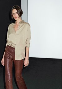 Camicia beige con bottoni e maniche arrotolate; abbinata a pantaloni in pelle marrone aderenti con una leggera svasatura all'orlo.