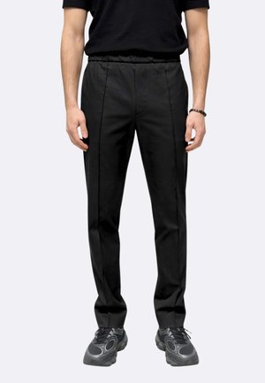 Uomo che indossa pantaloni neri slim fit con vita elastica, abbinati a scarpe da ginnastica nere e una maglietta nera, in piedi davanti a uno sfondo semplice.